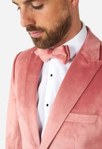 Chaqueta de esmoquin de terciopelo rosa con solapas muesca y un bolsillo. Acompañada de una pajarita a juego sobre una camisa blanca.