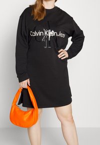 Černé oversized mikinové šaty s lesklým logem "Calvin Klein Jeans". V doprovodu hladké oranžové kabelky typu hobo.