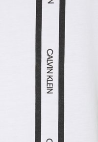 Bílý bavlněný tričko s černými vertikálními pruhy a opakujícím se textem "CALVIN KLEIN" pro branding. Hladká textura, klasický střih.
