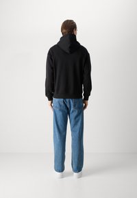 Felpa nera con un ampio cappuccio, indossata sopra jeans blu a taglio largo e scarpe da ginnastica bianche. L'outfit presenta un design casual con un look semplice e pulito.