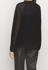 Femme portant un chemisier noir transparent à manches longues et un pantalon en cuir noir, vue de dos sur un fond uni.