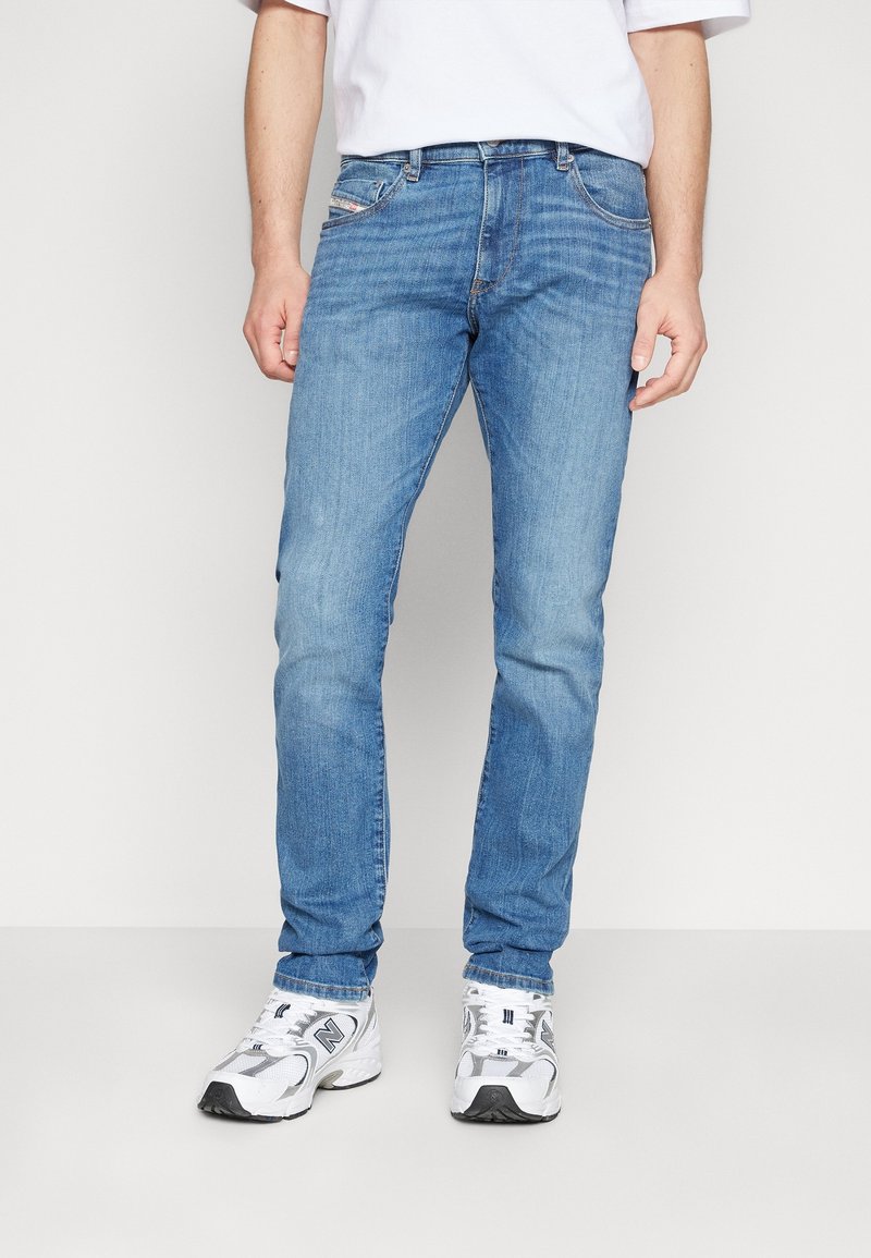 Personne portant un jean slim bleu clair, des baskets blanches et une chemise blanche à manches courtes, debout devant un fond blanc uni.
