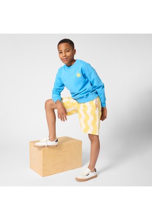 Αγόρι με μπλε φούτερ και κίτρινα σορτς με κυματιστό σχέδιο στέκεται με το ένα πόδι σε ξύλινο τούβλο σε απλό φόντο.