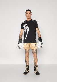 Schwarzes T-Shirt mit weißem Grafikdesign, goldene Satin-Shorts, schwarze Boxhandschuhe und schwarz-gelbe Schuhe. Tätowierungen an den Beinen sichtbar.
