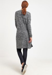 Długi szary i czarny kardigan z dzianiny o prążkowanej fakturze, z bocznymi rozcięciami i luźnym krojem, zestawiony z ciemnymi dżinsami i czarnymi trampkami.