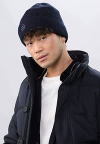 Navy knitted beanie with ribbed texture and a logo patch, worn with a black jacket and white shirt, showcasing a fitted design.