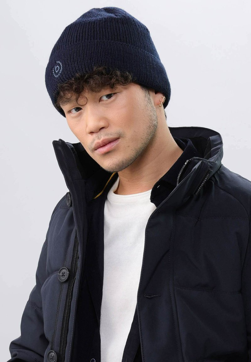Navy knitted beanie with ribbed texture and a logo patch, worn with a black jacket and white shirt, showcasing a fitted design.