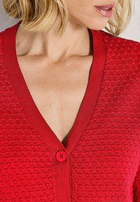 Eine Frau trägt einen strukturierten roten Cardigan mit Knöpfen und tiefem V-Ausschnitt, der einen Teil ihres Halses und ihrer Lippen zeigt.