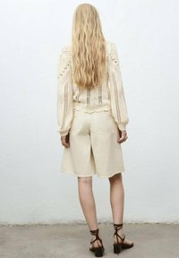 Gehaakte crèmekleurige trui met openworkpatroon, gecombineerd met lichtbeige shorts. Het model draagt bruine sandalen met bandjes en staat voor een simpele lichtgekleurde achtergrond.