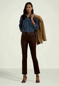 Femeie îmbrăcată într-o cămașă din denim, pantaloni din piele maro închis și sandale cu toc înalt, ținând o jachetă cu carouri maro peste umăr.