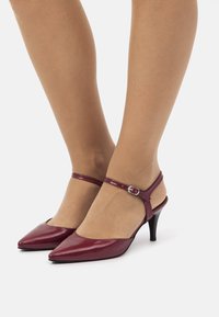 Tacones de charol burdeos con punta afilada, tira delgada en el tobillo y un tacón de aguja de 5 cm. Textura suave, hardware mínimo.