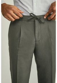 Šedé plisované kalhoty s širokou šňůrkou v pase, vyrobené z hladké tkaniny. Mají střední přední šev a vertikální kapsy.