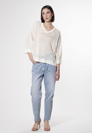 Leichter, cremefarbener Netzpullover mit V-Ausschnitt und lockerer Passform, kombiniert mit hellblauen, geraden Jeans und beigen Absätzen.