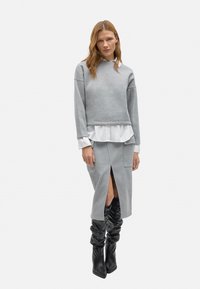 Sudadera gris corta superpuesta sobre una camisa blanca con dobladillo con volantes, combinada con una falda gris con una abertura frontal y botas negras hasta la rodilla.