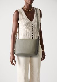 Borsa a spalla verde in pelle con una texture liscia e hardware in argento, indossata a tracolla con un design minimalista. Presenta una tracolla regolabile.