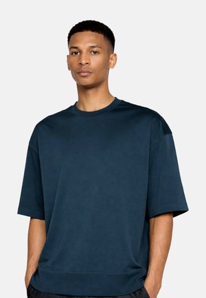 Uomo con capelli corti che indossa una maglietta ampia blu scuro con maniche corte e collo rotondo, in piedi con le mani in tasca su uno sfondo semplice.