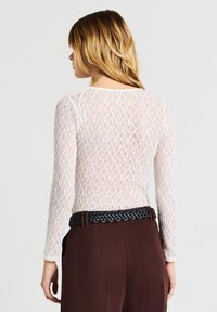 Top bianco in pizzo a maniche lunghe con un motivo delicato, abbinato a pantaloni neri a vita alta di colore marrone scuro e una cintura nera intrecciata.