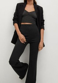 Vrouw in een donker gestreepte blazer, een kort gebreid tanktop en hoge, uitlopende donkere broek tegen een effen achtergrond.