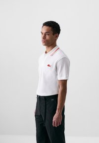 Chemise polo blanche avec un col à rayures orange, logo Lacoste en rouge sur la poitrine. Associée à un pantalon noir avec cordon de serrage et accent de logo.