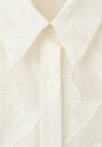 Chemise blanche à col avec un motif floral texturé subtil et des boutons blancs sur la patte de boutonnage avant.