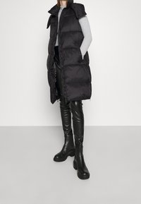 Schwarzige gepolsterte ärmellose Weste mit hohem Kragen, seitlichen Taschen und glänzender Textur, getragen über einem grauen Pullover und schwarzen Lederhosen.