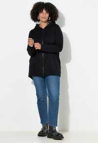 Mujer con cabello rizado que lleva pendientes de aro, sudadera negra con cremallera, jeans azules y botas negras gruesas, de pie contra una pared blanca.