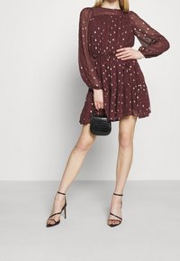 Burgundy klänning med långa, puffärmar, prydd med gyllene prickar och en vågig kjol. Matchas med en liten svart handväska i krokodilsmönster.