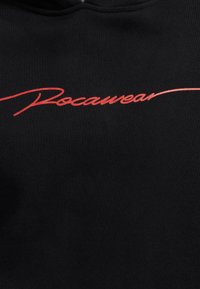 Svart hettegenser med rød kursiv tekst "Rocawear." Laget av mykt stoff med ribbet tekstur og standard hettegensdesign.