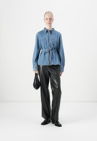 Lichtblauwe denim shirt met een strik in de taille, voorzien van twee borstzakken. Gepaard met zwarte leren broek en enkellaarsjes, houdt een zwarte tas vast.