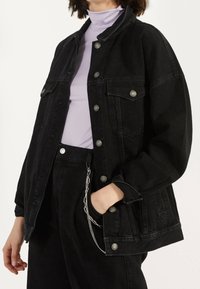 Persona vestida con chaqueta y pantalones de mezclilla negros con botones plateados y cadena, combinados con un suéter de cuello alto lila claro.