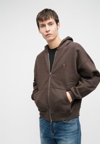 Pruun tõmbluku kapuutsiga särk, millel on ees tõmblukk ja kaks küljetaskut. Pehme materjal, lõtv istuvus ja väike broderie logo vasakul rinnal.