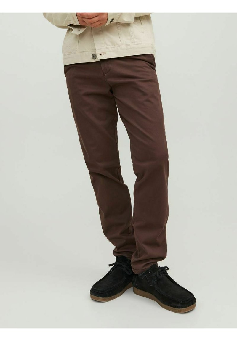 Pantaloni in cotone marroni con un design slim fit, abbinati a scarpe in suede nere. Il modello indossa un giubbotto chiaro e si trova davanti a uno sfondo neutro.