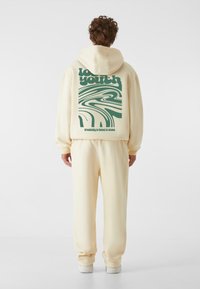 Sudadera color crema con un gráfico verde audaz en la parte posterior, combinada con pantalones de chándal crema a juego y zapatillas blancas. Textura suave.