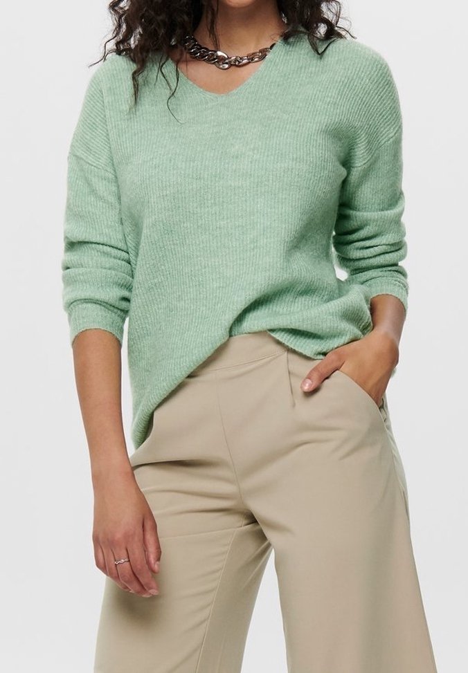 Pull vert clair à col en V avec texture côtelée, associé à un pantalon beige. Il présente des manches longues et une coupe décontractée, accompagné d'un collier en chaîne argentée.