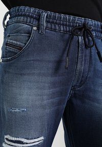 Pantalones jogger de mezclilla de color azul oscuro, con cintura elástica y cordón. Presentan áreas desgastadas en las rodillas y un diseño de cinco bolsillos.