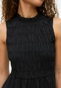 Robe noire sans manches avec un col haut à volants et une texture froncée sur le corsage, se prolongeant par une jupe lisse.