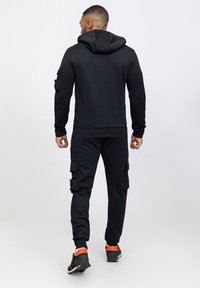 Sweat à capuche noir en tissu texturé, associé à un pantalon cargo assorti avec poches, complété par des baskets.
