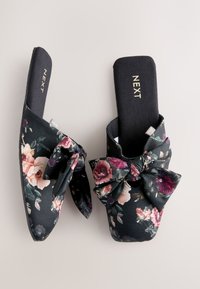 Mules con estampado floral sobre un fondo negro, con un gran lazo en cada zapato, fabricados con tela texturizada y suela plana.