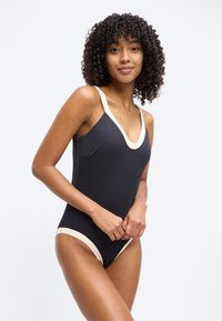 Γυναίκα με σγουρά μαλλιά που φοράει μαύρο ολόσωμο μαγιό με λευκή επένδυση, στέκεται με τα χέρια δεμένα, μπροστά σε ένα απλό φωτεινό φόντο.