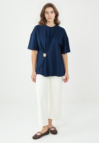 Navy-blaues T-Shirt mit kurzen Ärmeln und einem gedrehten Design, verziert mit einem goldenen Knopf, kombiniert mit weiten weißen Hosen und braunen geflochtenen Ballerinas.