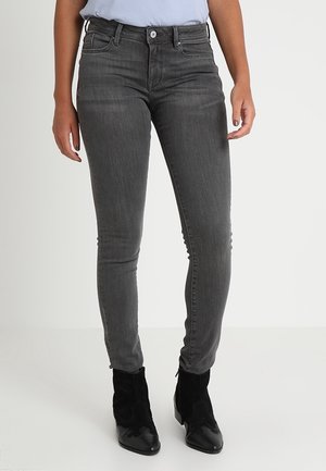 Person, der dunkelgraue, eng anliegende Jeans und schwarze Stiefeletten trägt und vor einem schlichten, hellen Hintergrund steht.