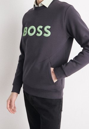 Osoba nosící tmavý svetr s mintově zeleným nápisem "BOSS", pod ním bílou košili s límečkem a jednou rukou v přední kapse.
