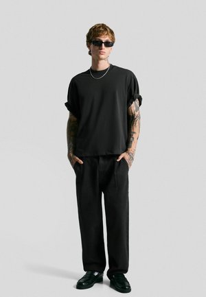 Black short-sleeve shirt with rolled cuffs, paired with loose black trousers and black shoes. Accessories include silver chain and sunglasses.