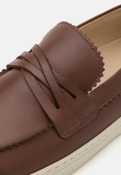 Chaussure slip-on en cuir marron avec une surface texturée, des accents festonnés et une semelle en caoutchouc blanc. Présente un détail en cuir entrelacé à l'avant.