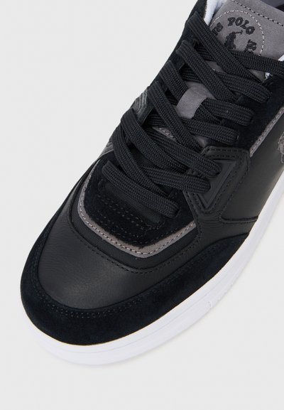 Gros plan sur une sneaker noire avec une tige en suède et en cuir, ornée d'accents gris et de lacets noirs, sur fond uni.
