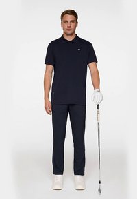 Marinblå pikétröja med korta ärmar och krage; matchande marinblå byxor; person som håller i en golfklubba; vita sneakers; slät tyg, minimalistisk design.