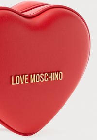 Borsa a forma di cuore rossa realizzata in materiale sintetico testurizzato, caratterizzata da un logo dorato "LOVE MOSCHINO" sulla parte anteriore. Bordo e curve lisci.