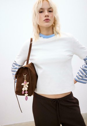 Junge Frau mit blonden Haaren trägt ein weißes, bauchfreies Oberteil über blau gestreiften Langarmshirts, dunkle Hosen und trägt eine braune Wildledertasche mit rosa Perlenanhänger.