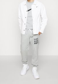 Person med vit jacka, grå Nike-t-shirt, grå Nike-träningsbyxor med text på vänster ben och vita Nike höghänstens sneakers.