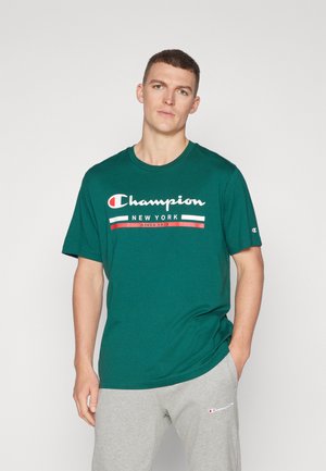 Camiseta de algodón color turquesa con el logo "Champion" en blanco y rojo, diseño de cuello redondo, mangas cortas, combinada con pantalones de chándal grises.